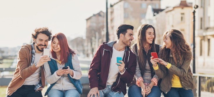 5 Personen sitzen nebeneinander und zeigen sich gegenseitig etwas auf ihren Smartphones