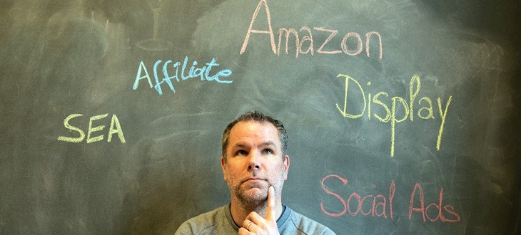 Mann sitzt vor Tafel mit den Aufschriften SEA Affiliate Amazon Display und Social Ads